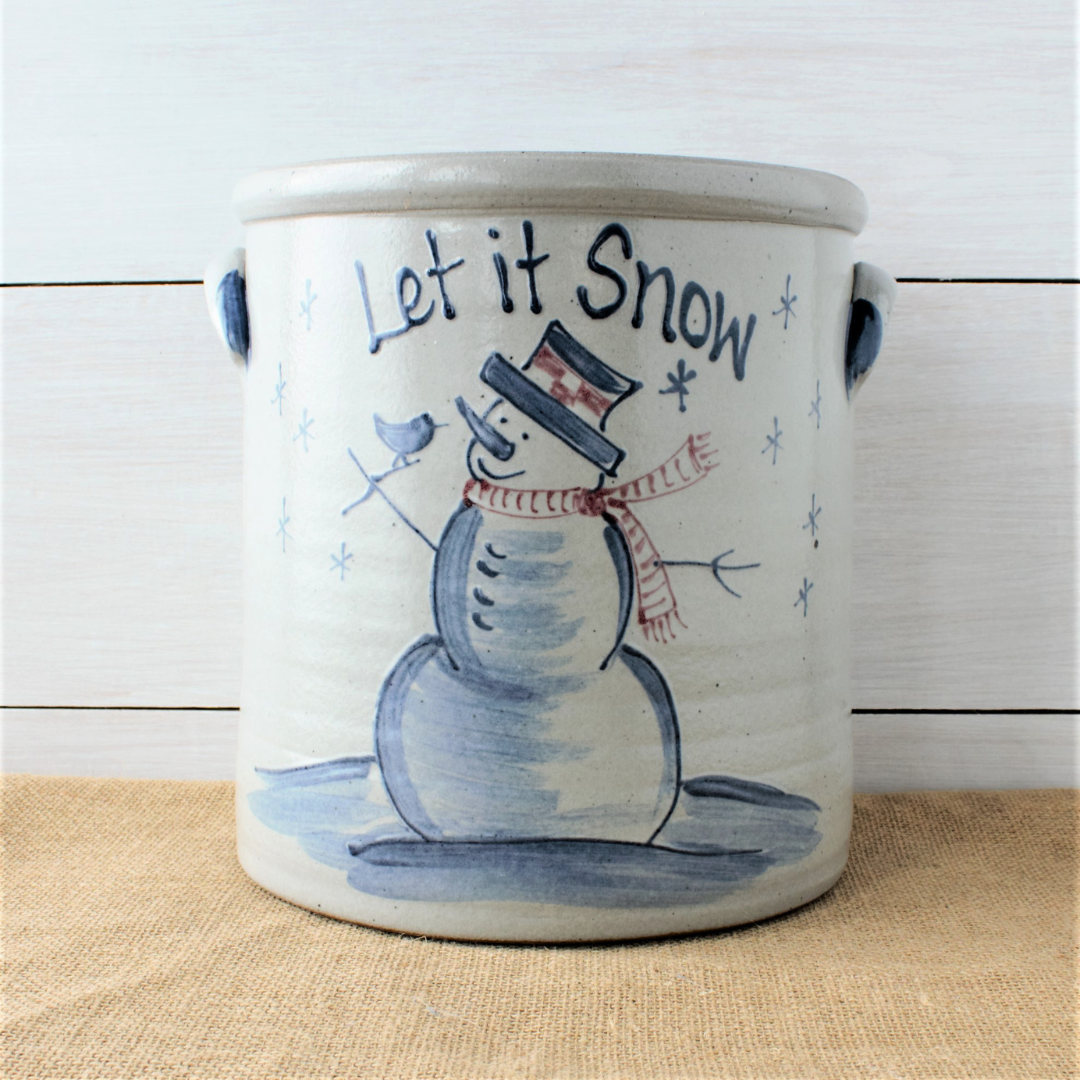Hand thrown 2 gallon crock with a hand decorated snowman pattern, snowman has a hat and scarf and small bird in hand, the words Let It Snow are painted above the design